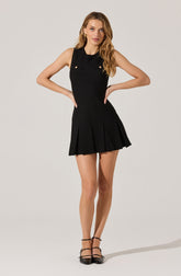 A woman is wearing a sleeveless black dress with a fitted bodice. The dress has decorative gold buttons on the chest area and features a pleated skirt that flares out slightly. She is also wearing black strappy high-heeled shoes.